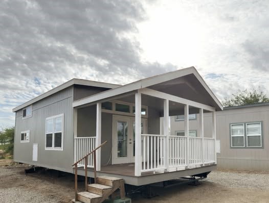 A small, modern gray house with a white porch is set under a cloudy sky. The house has double glass doors and several windows, creating a serene ambiance.