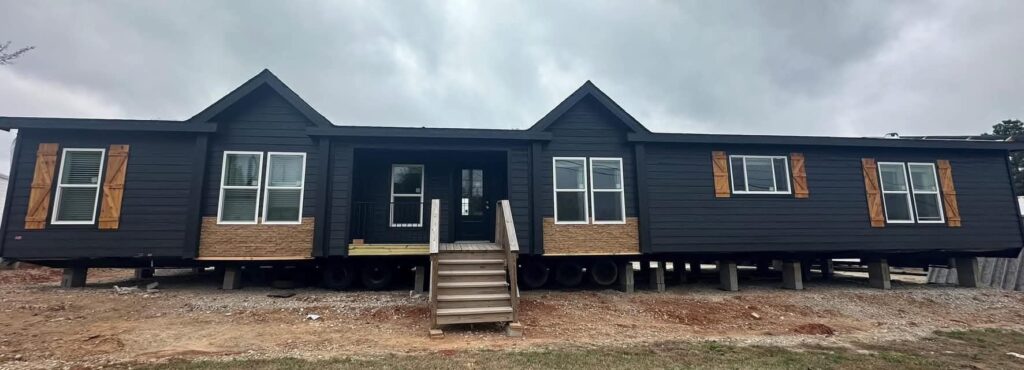Wide, dark-colored mobile home with stone accents, white-trimmed windows, and wooden shutters. Wooden steps lead to the front door under a cloudy sky.