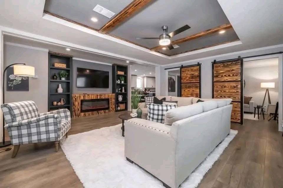 Stylish living room with a white sofa, plaid armchair, and rustic wood accents. Barn doors, ceiling beams, and modern fan create a cozy, elegant vibe.