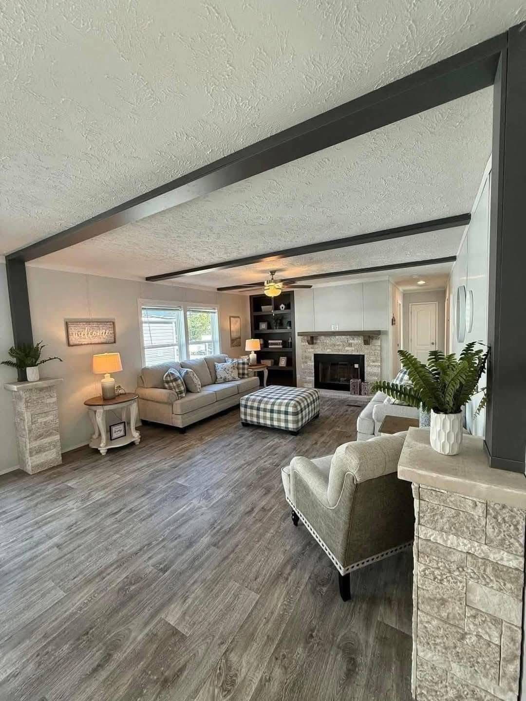 Spacious living room with light wood floors, a beige sofa, plaid ottoman, and armchair. A fireplace is centered, surrounded by shelves. Warm, cozy ambiance.