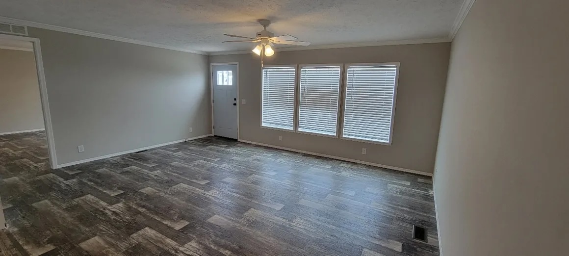 A spacious, empty living room with gray walls, wooden flooring, and large windows with blinds. A ceiling fan with lights hangs in the center. Quiet and inviting.