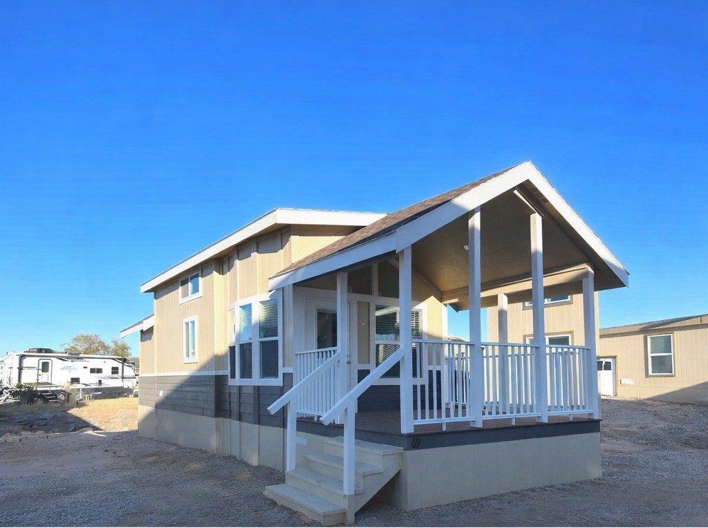 A small beige modular home with a spacious porch and white railing sits under a bright blue sky. The setting is sunny and open, conveying simplicity.