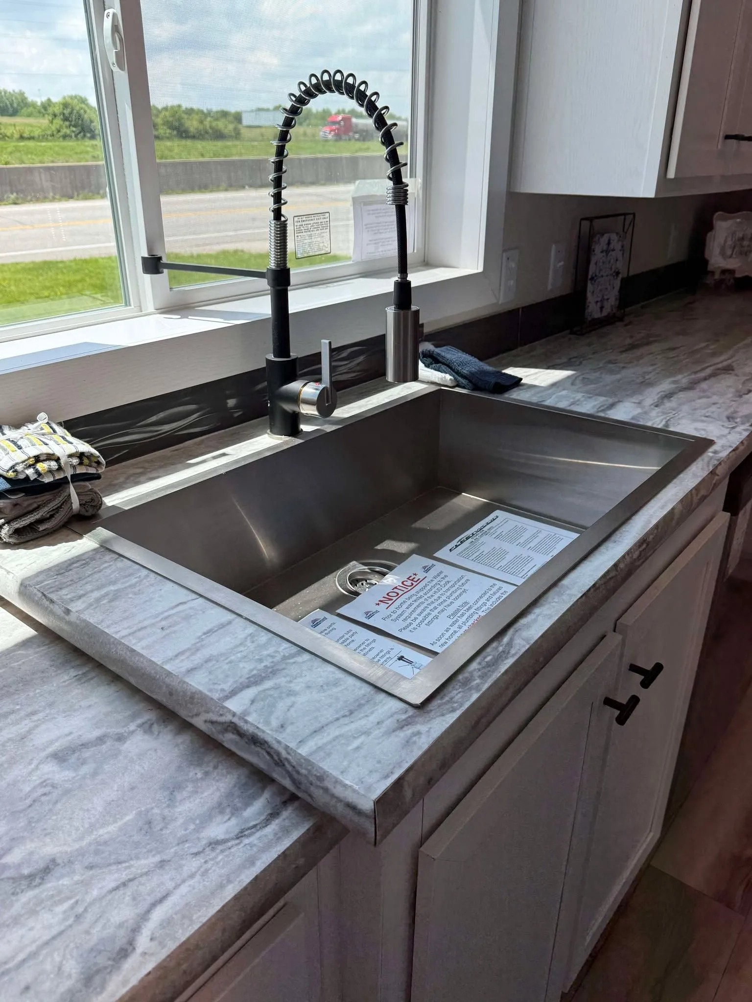 Sleek kitchen sink with a high-arched faucet on a marble countertop, next to a large window with a view of a sunny road and greenery.
