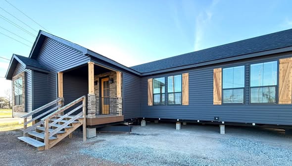 A modern manufactured home with dark gray siding and wooden shutters. A small porch with stone accents and stairs leads to the entrance under a clear blue sky.
