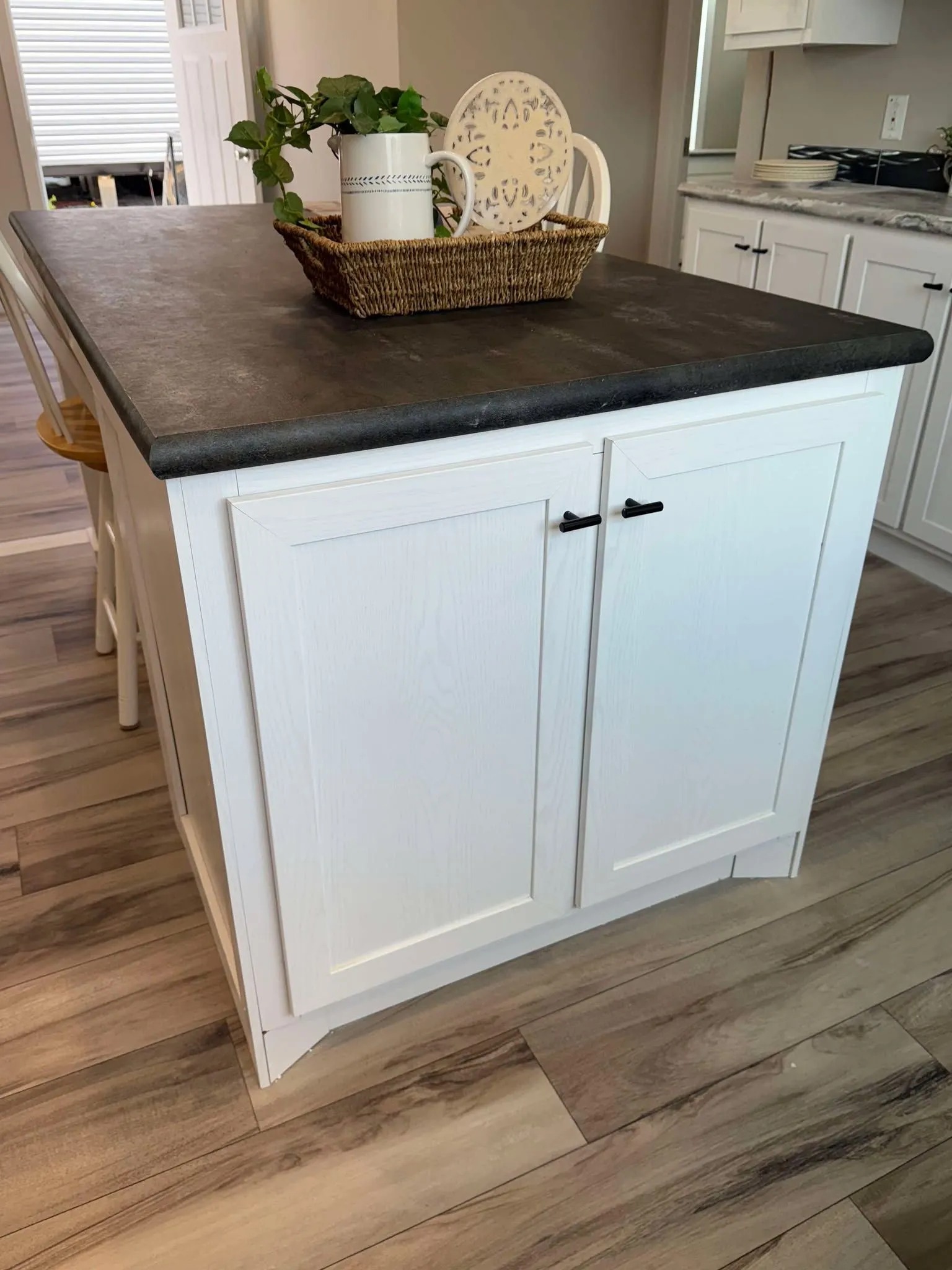 A kitchen island with a dark countertop and white cabinets stands on wooden flooring. A woven basket with a plant and decorative plates sits on top, creating a cozy, inviting atmosphere.