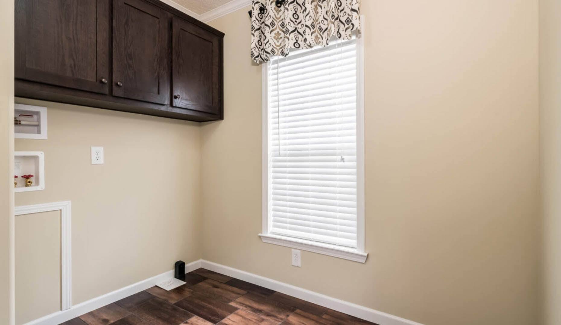 A small room with beige walls and dark wood flooring features a large window with blinds and a patterned valance. Dark wood cabinets are mounted above.