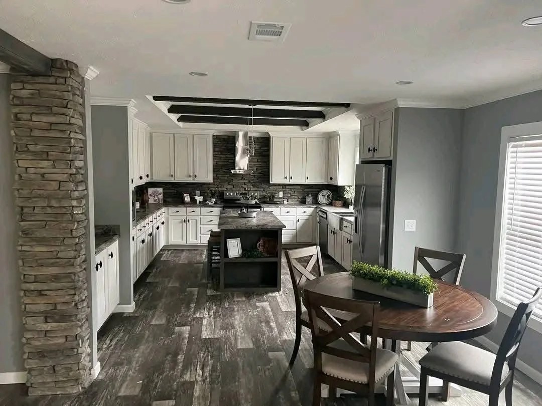 Modern kitchen with white cabinets, stone backsplash, and a central island. A round table with chairs and greenery adds warmth. Natural light streams in.
