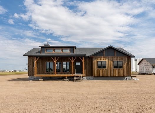 A rustic wooden cabin with large windows and a covered porch sits on a gravel area under a partly cloudy sky, evoking a tranquil rural atmosphere.