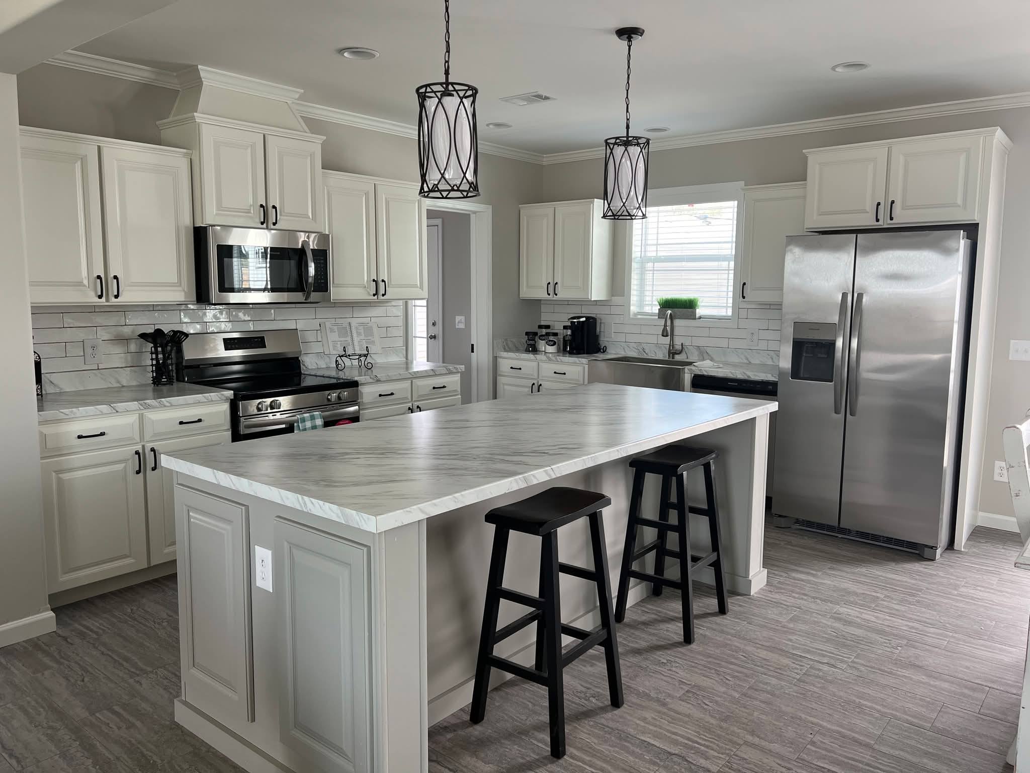 Modern kitchen with white cabinets, stainless steel appliances, and a large marble island with two black stools. Two pendant lights hang above.