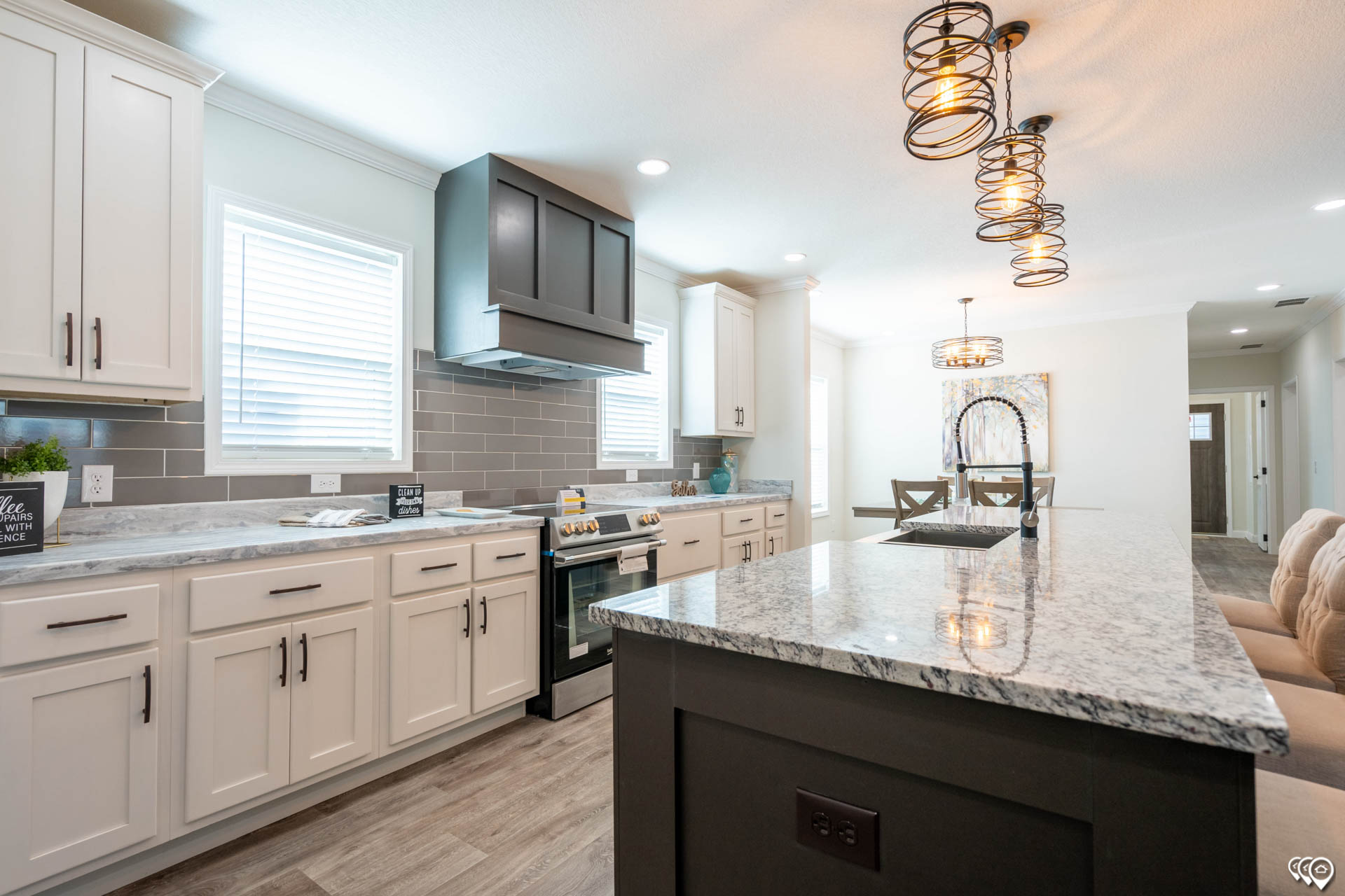 Modern kitchen with light wood flooring and white cabinets. A gray granite island with black accents is centered, under spiral pendant lights. Bright and inviting atmosphere.