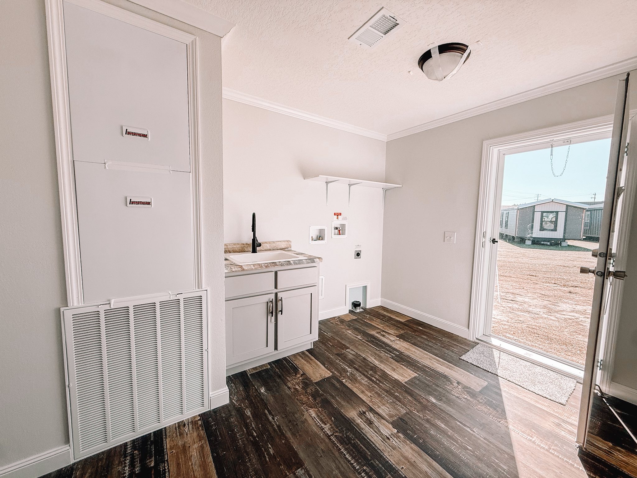 Bright laundry room with wood flooring, white cabinets, and a countertop. An open door leads outside to a sunny view of mobile homes. Calm and airy ambiance.