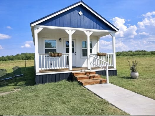 A quaint tiny house with blue and white siding sits on a grassy field under a bright blue sky. The porch features a white railing and hanging baskets.