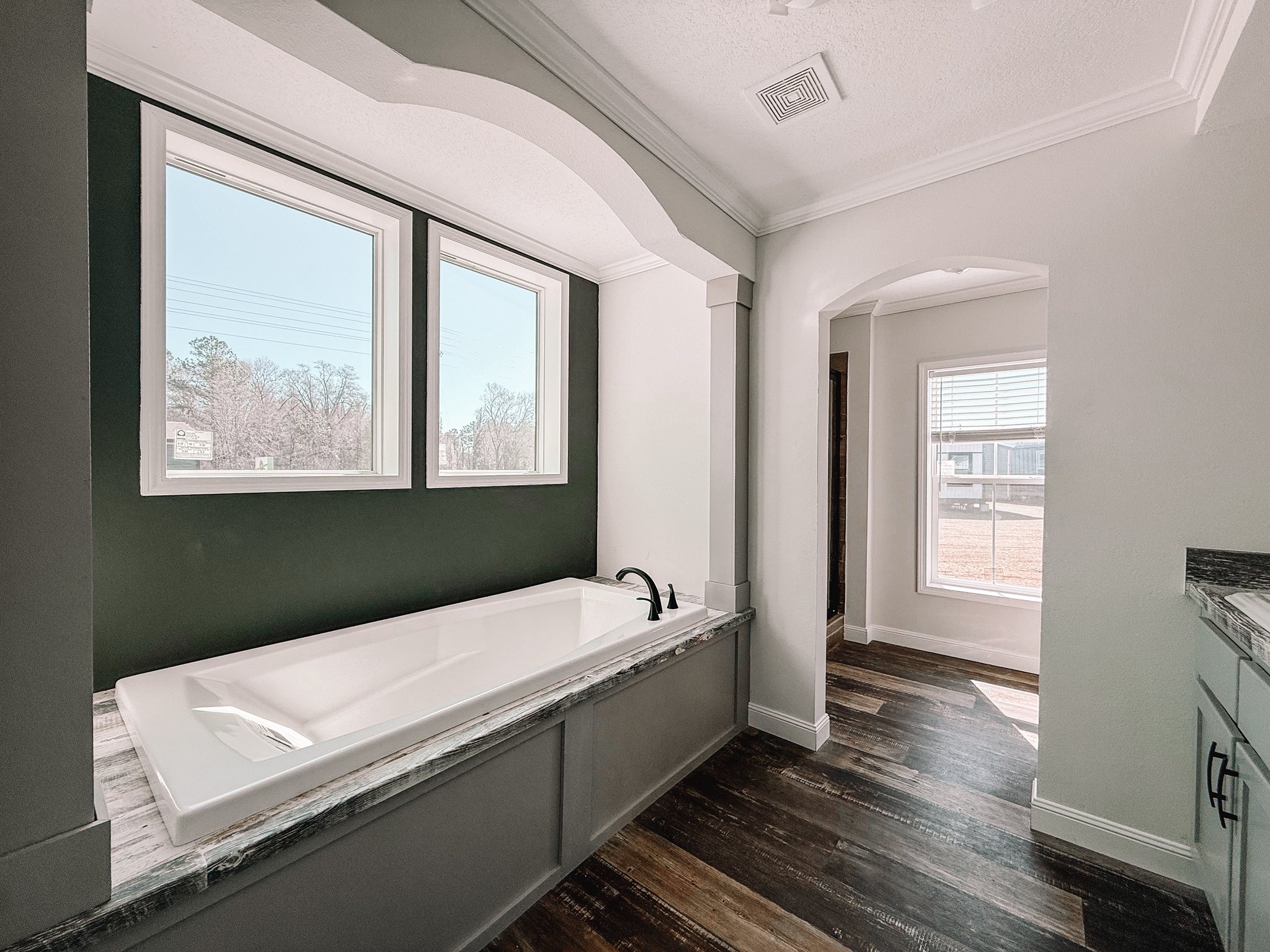 Luxurious bathroom featuring a large white bathtub with a dark green accent wall and two windows. Hardwood floors and an arched doorway create an elegant look.