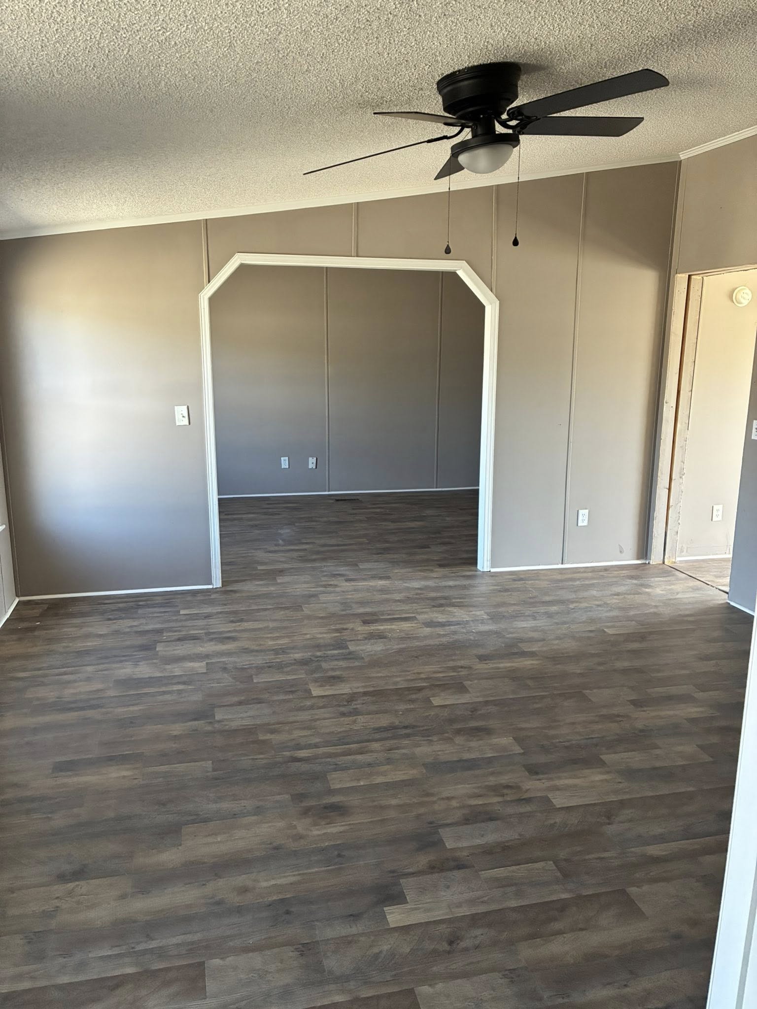 Empty room with wooden laminate flooring, beige walls, and a white ceiling. A black ceiling fan is centered above. Archway leads to another room.