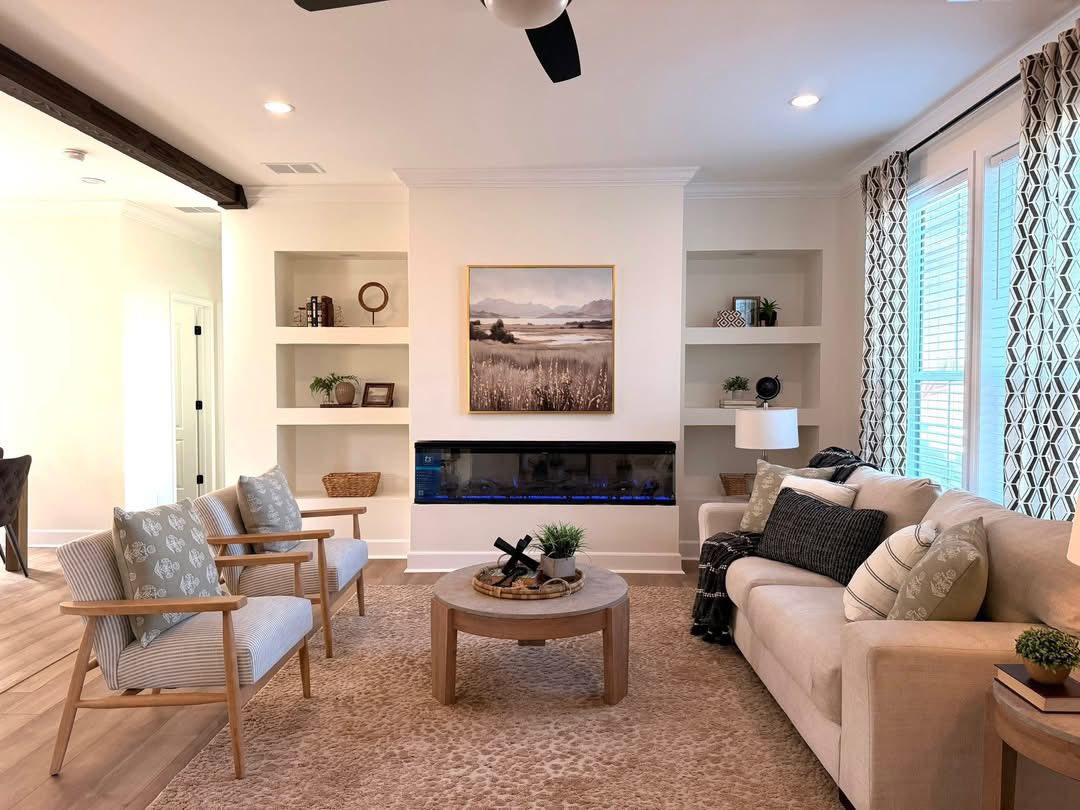 Cozy living room with beige sofa, patterned cushions, two armchairs, and a round wooden table. Modern fireplace and nature painting add warmth.