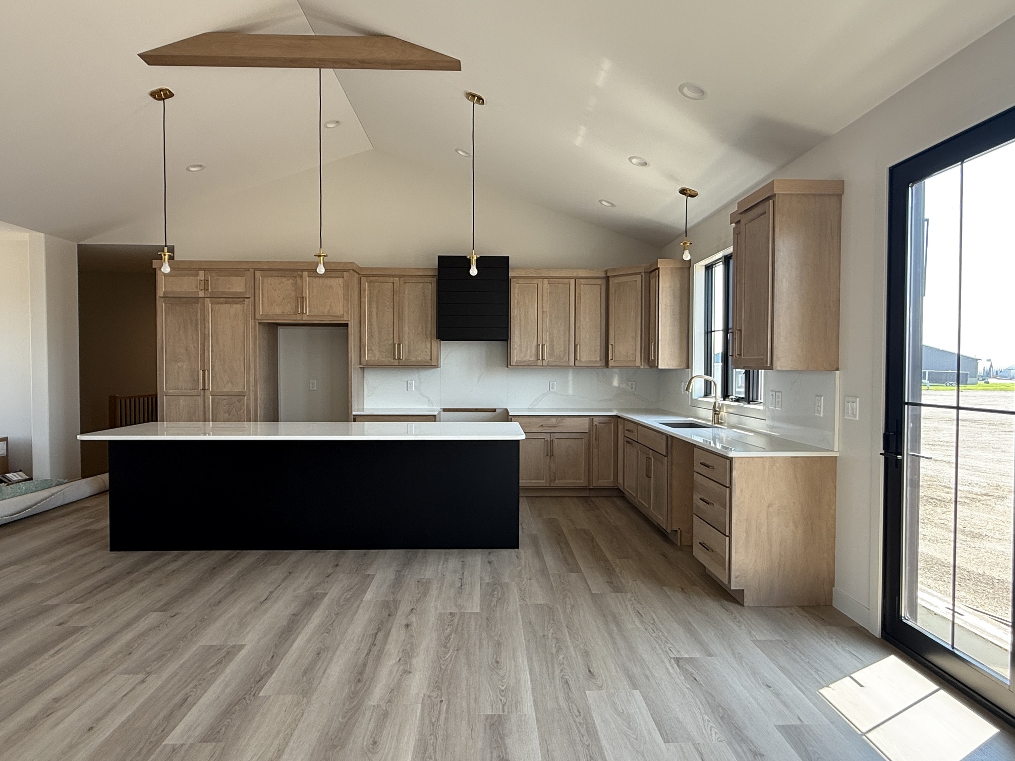 Spacious modern kitchen with light wood cabinets, a large black island, and wood flooring. Natural light streams in through large windows, creating an airy feel.