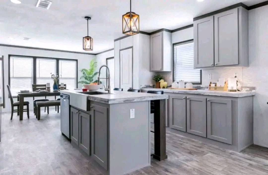 Modern kitchen with gray cabinets, an island with a marble countertop, and pendant lights. A dining table is in the background with a potted plant. Bright, airy atmosphere.