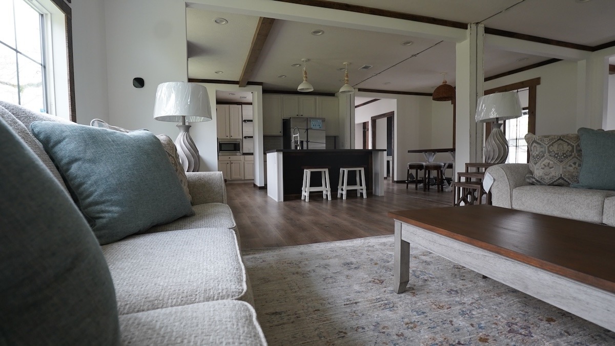 Cozy living room with a light gray sofa, soft blue cushions, and a wooden coffee table. Open kitchen with a black island and stools in the background.