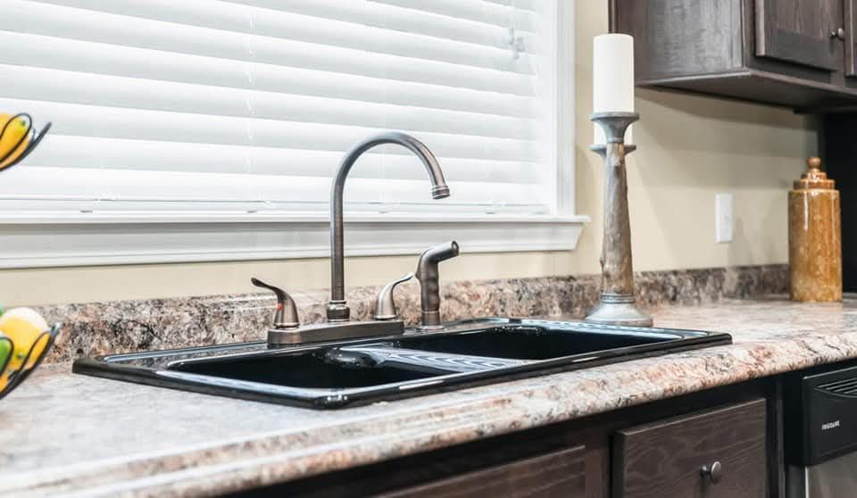 Modern kitchen sink with a sleek stainless steel faucet, set in marble countertops. Wooden cabinets, a tall candle, and fruit bowl add warmth. Clean, elegant ambiance.