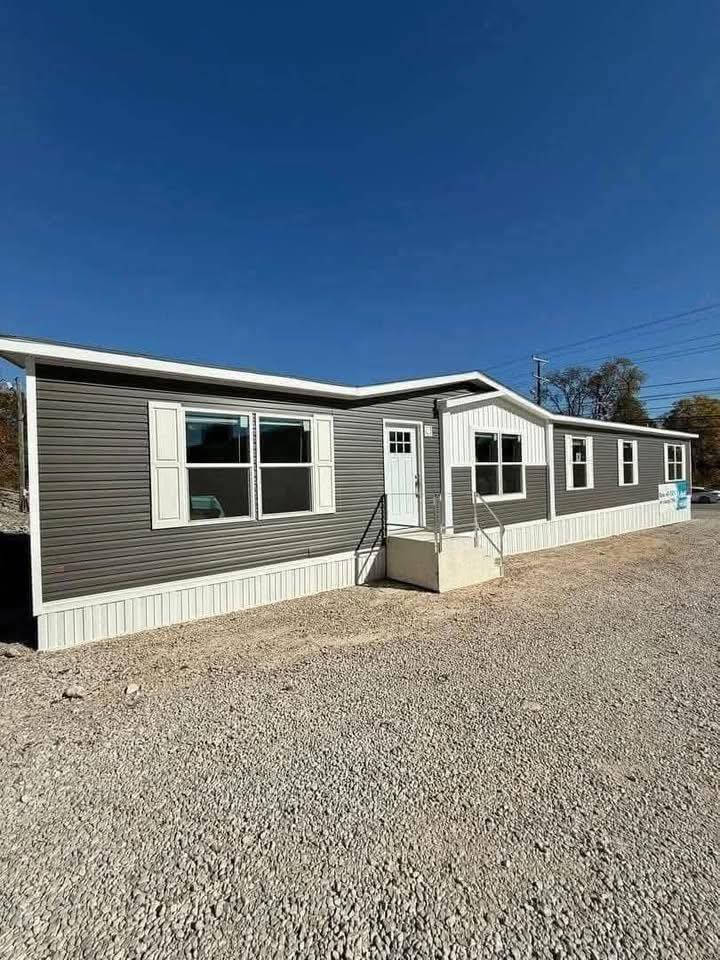 Single-story gray modular home with multiple windows and a central white door on a gravel lot. The sky is clear and blue, creating a calm atmosphere.
