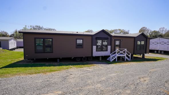 A brown modular home with white steps leading to the entrance sits on a gravel area surrounded by green grass under a clear blue sky.