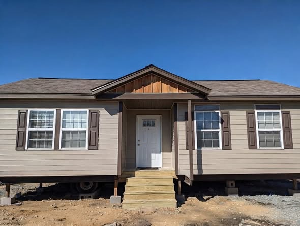 A beige modular house with a gabled roof, two front windows with shutters, and a central white door. It sits elevated on concrete blocks under a clear blue sky.