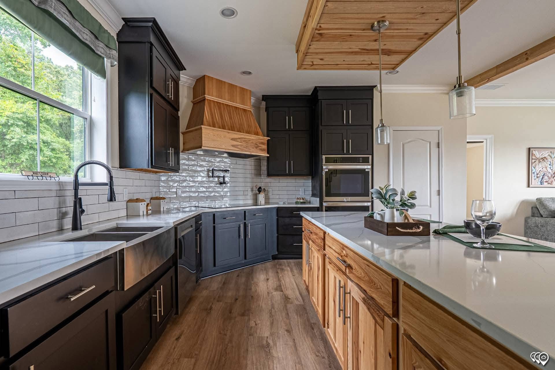 Modern kitchen with dark cabinets, white countertops, and wood accents. Features a farmhouse sink, brick backsplash, and natural light from a large window.