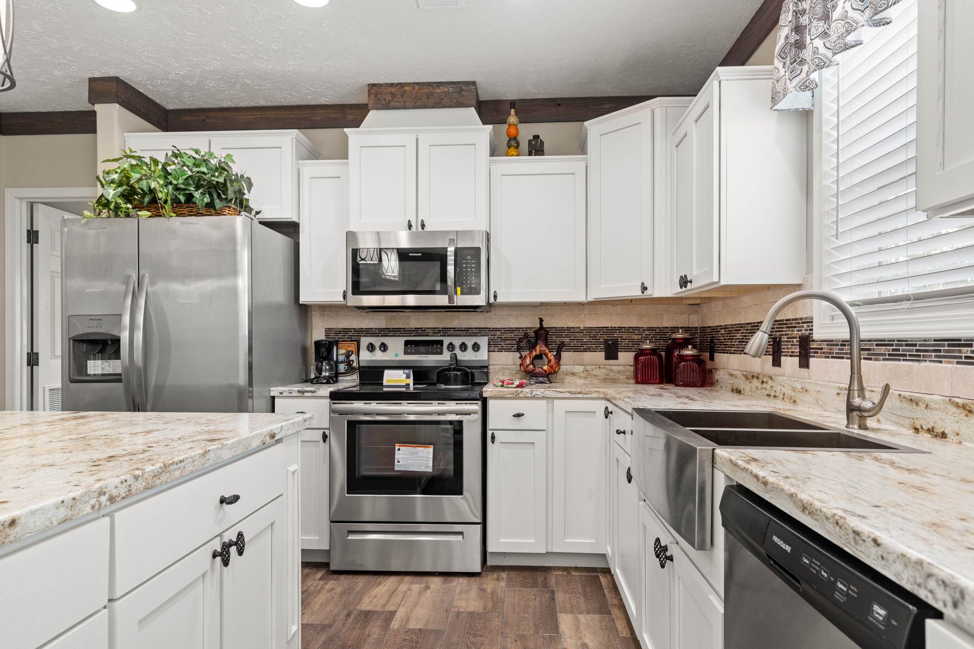 A modern kitchen with white cabinets, stainless steel appliances, and a marble countertop. Includes plants atop the fridge and wooden flooring for warmth.