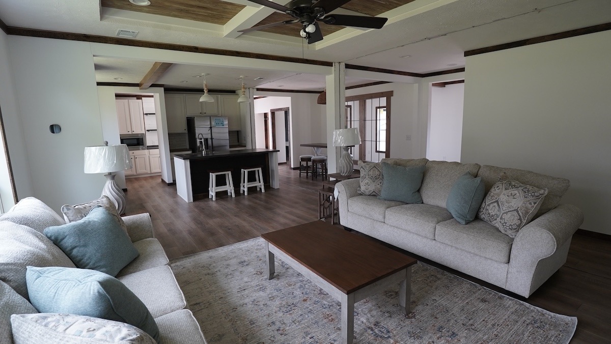 A cozy, modern living room featuring two beige couches with blue and patterned pillows, a wooden coffee table, and an adjacent open kitchen with an island. The space is inviting and well-lit with natural light, dark wood floors, and a ceiling fan.