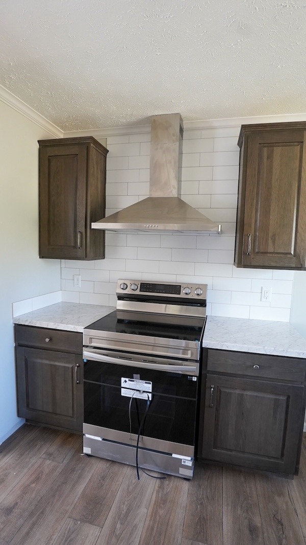 Modern kitchen with stainless steel stove and vent hood, surrounded by brown wood cabinets and white tile backsplash. The floor is light wood.