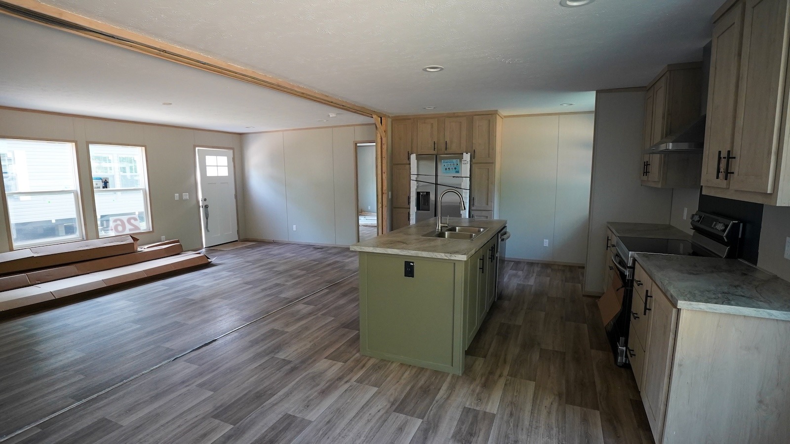 Spacious open-plan kitchen and living area with wood flooring. Features green island with sink, stainless steel appliances, light wood cabinets.