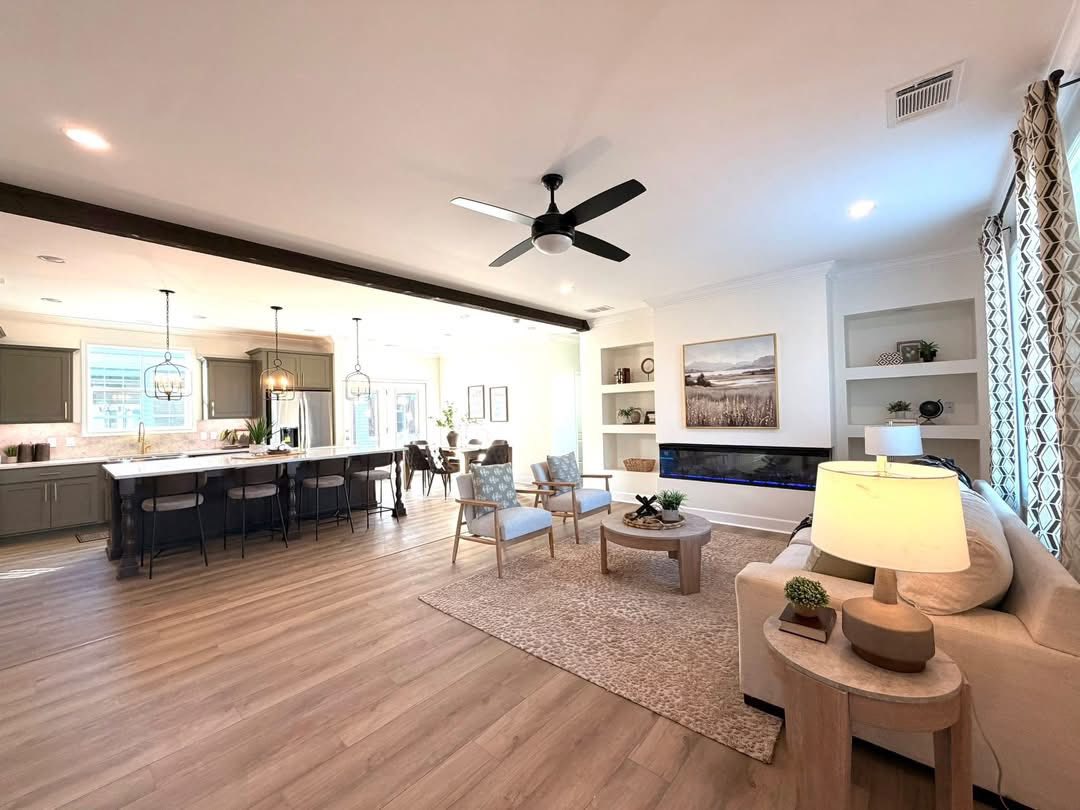 Open-concept living room with light wood floors, a beige sofa, and two armchairs on a rug. Kitchen and dining area visible, featuring pendant lights. Airy and modern.