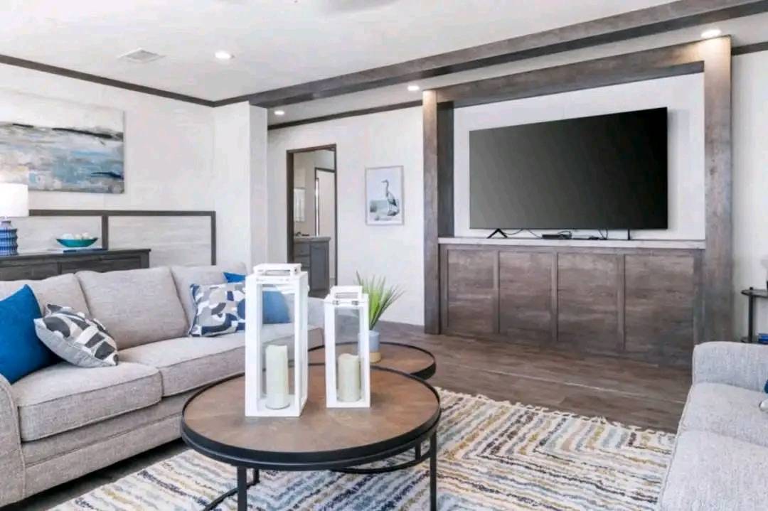 Modern living room with neutral tones, featuring a large flat-screen TV on a wooden stand. Two beige sofas with blue and patterned cushions face a round coffee table holding decorative lanterns and a small plant. Abstract art and a rug add a cozy feel.