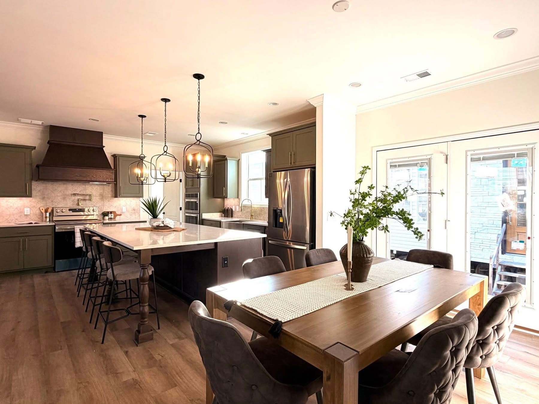 Spacious kitchen with wood floors, olive cabinets, and a large island with stools. A dining table with chairs and a vase with green branches. Bright, cozy atmosphere.
