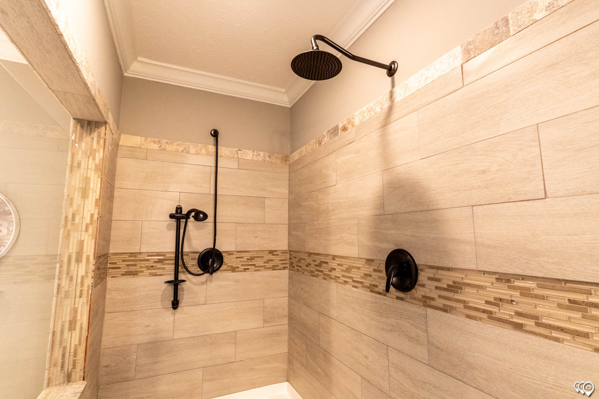 Elegant shower with beige tile walls, featuring a black rain showerhead and handheld shower. A mosaic accent stripe adds a stylish touch.