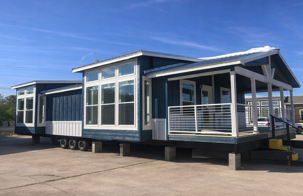 A modern tiny house on wheels with a blue exterior and large windows. It has a covered porch with white railings and a metal roof, situated on a sunny day.