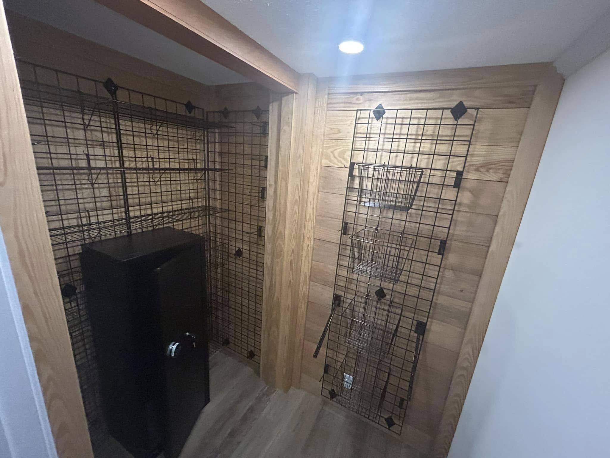 A small room with wooden walls and metal grid panels. A black safe stands in the corner, lit by a ceiling spotlight. The ambiance is utilitarian and secure.