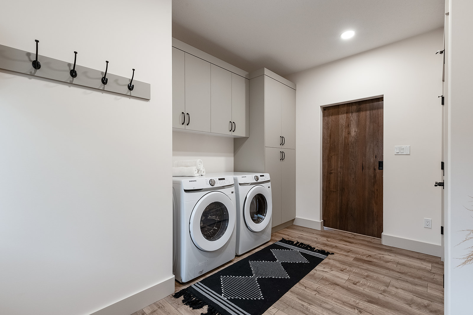 A bright, modern laundry room features a white washer and dryer set, gray cabinets, wood flooring, a geometric rug, and wall hooks. Cozy and organized.