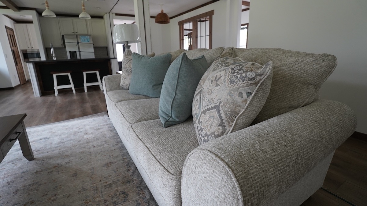 Beige sofa with patterned and teal pillows in a cozy living room, adjacent to a kitchen with pendant lights, wooden floors, and a homely ambiance.