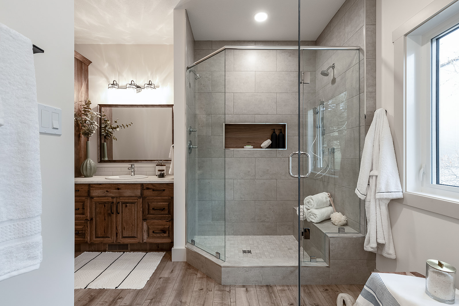 Modern bathroom featuring a glass-enclosed shower with gray tiles, wooden vanity, large mirror, and soft lighting. Towels and decor add a cozy feel.
