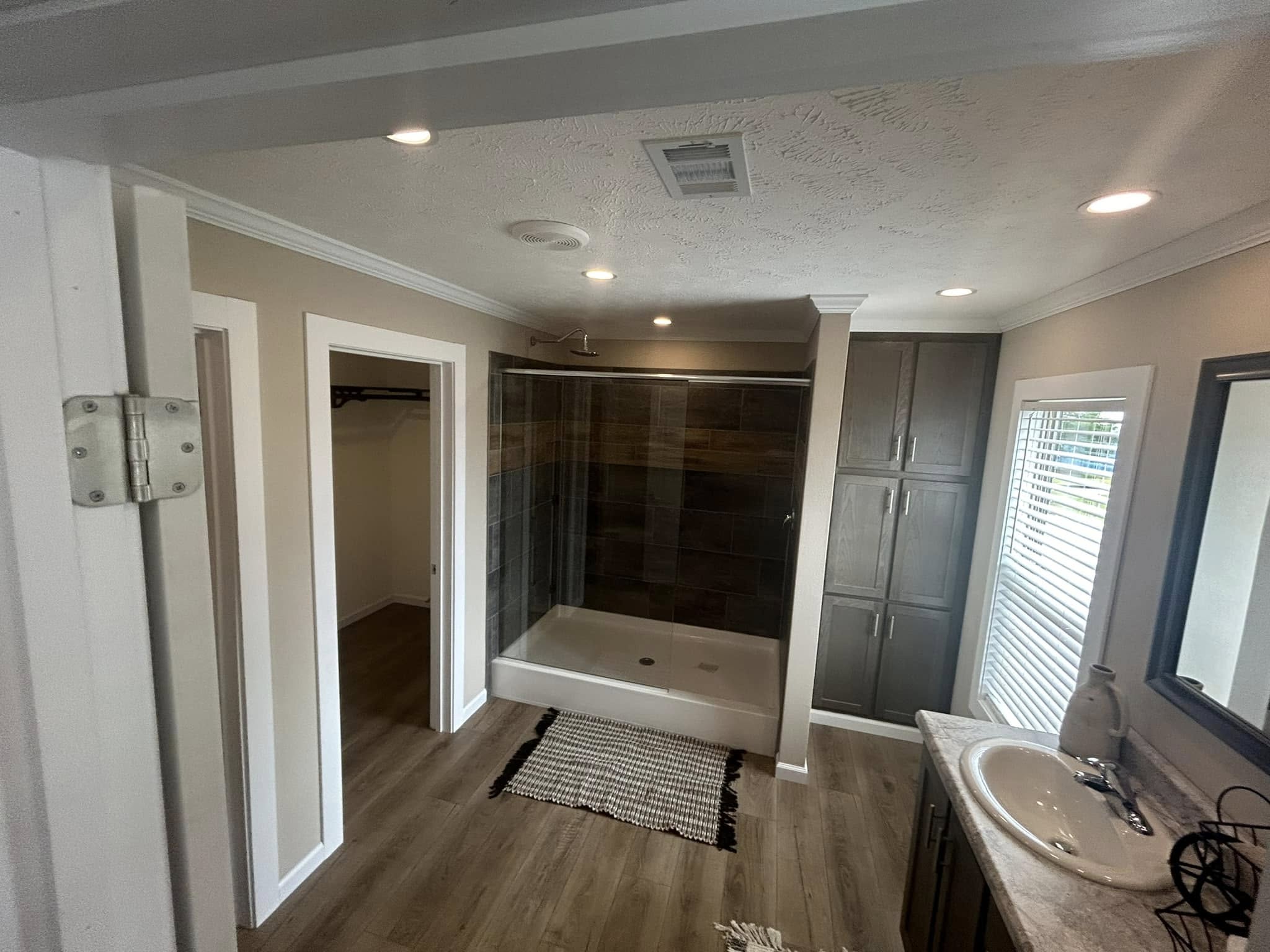 Spacious bathroom with wood flooring, large shower with glass doors, storage cabinets, sink with a mirror, and soft lighting creating a cozy ambiance.