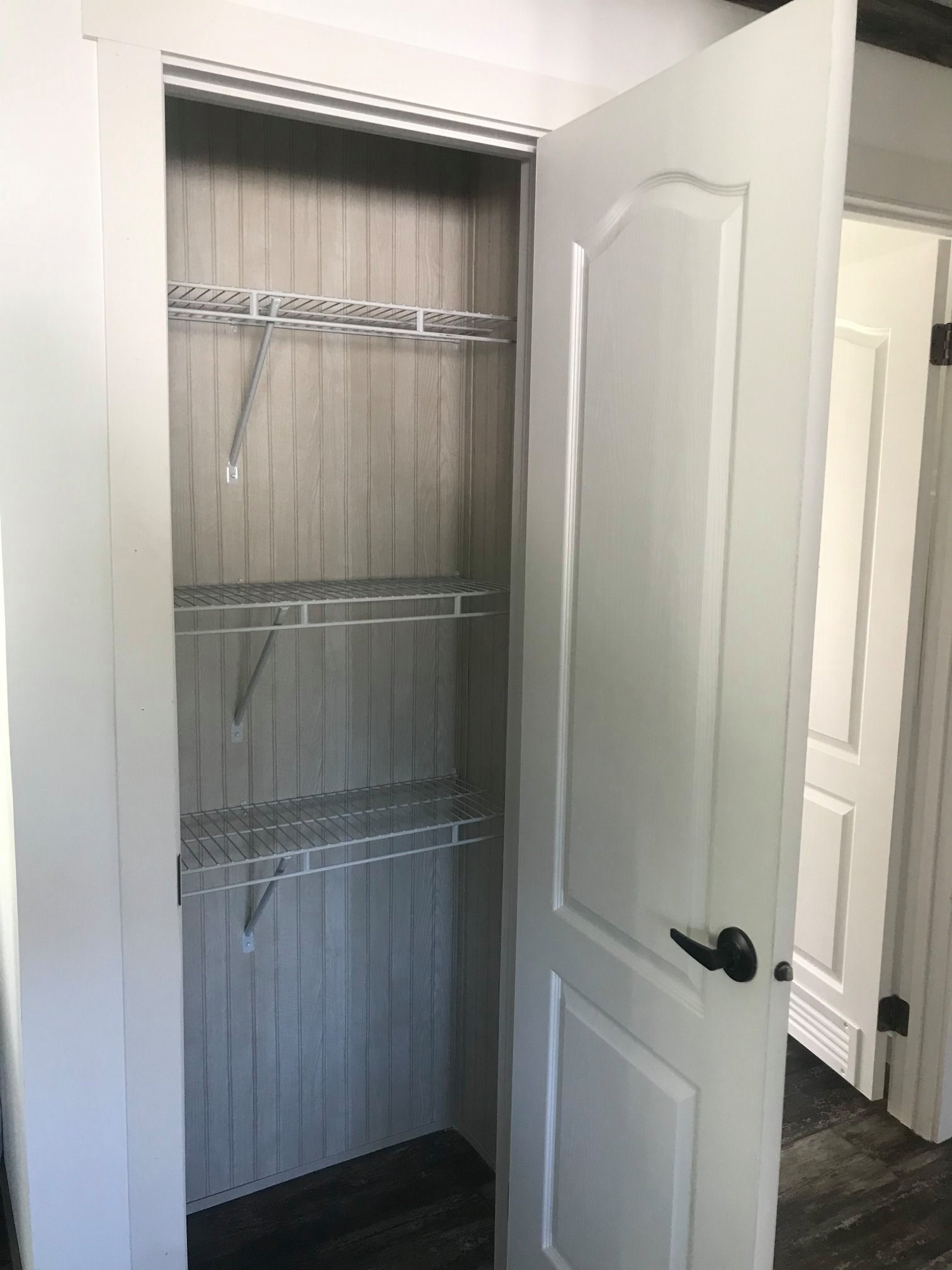White closet with an open door reveals three empty wire shelves against a light wood panelled back, creating a minimalist and organized appearance.