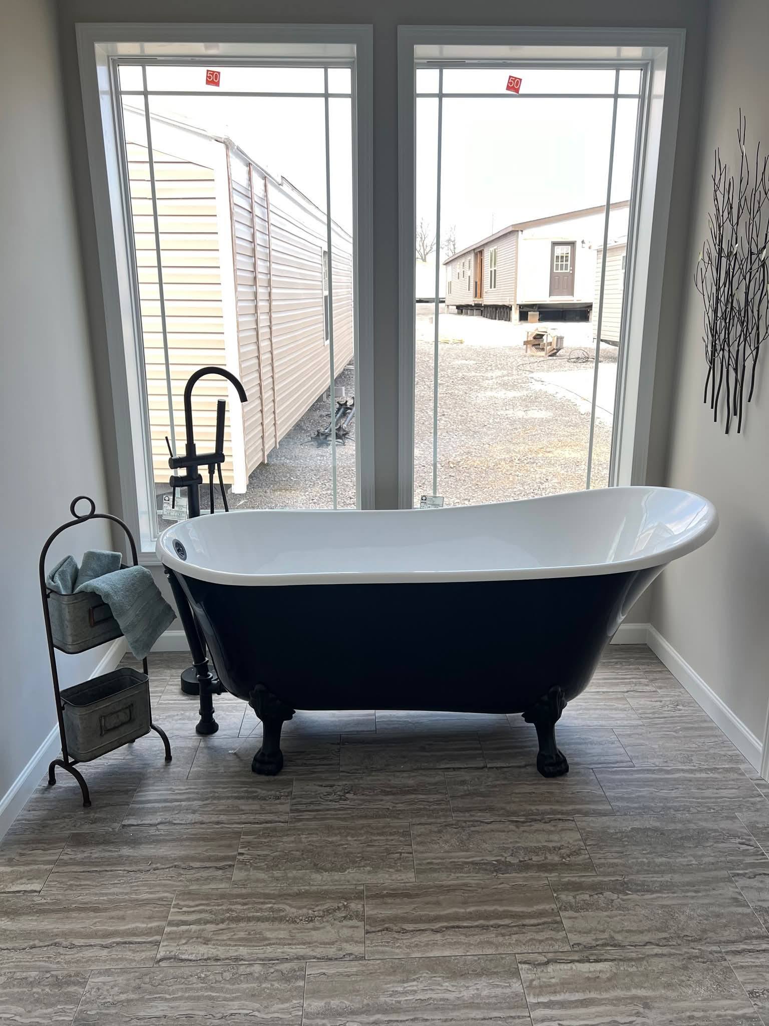 Freestanding clawfoot bathtub in a bright room with two large windows, beige tiled floor. Towel rack nearby, outside view of mobile homes.