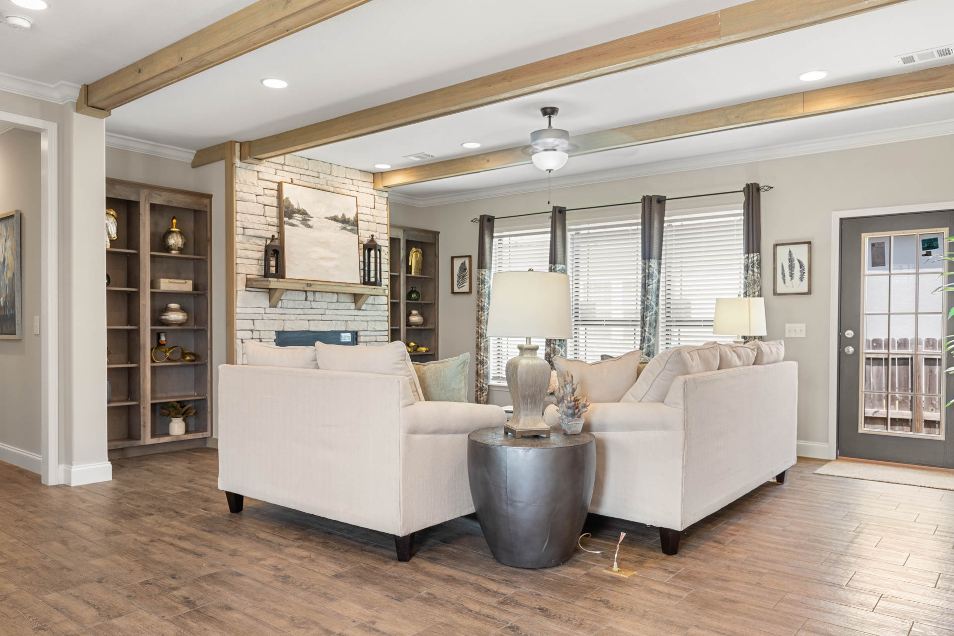 Spacious living room with beige sectional, decorative cushions, and two lamps. Stone fireplace, wooden beams, and large windows create a cozy ambiance.
