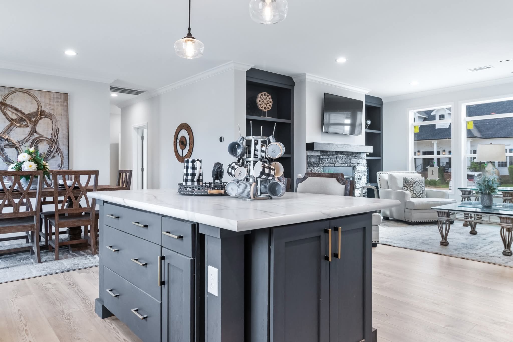 Modern open-plan living space with dark kitchen island, light countertops, elegant dining area, cozy living room with fireplace, and large windows.