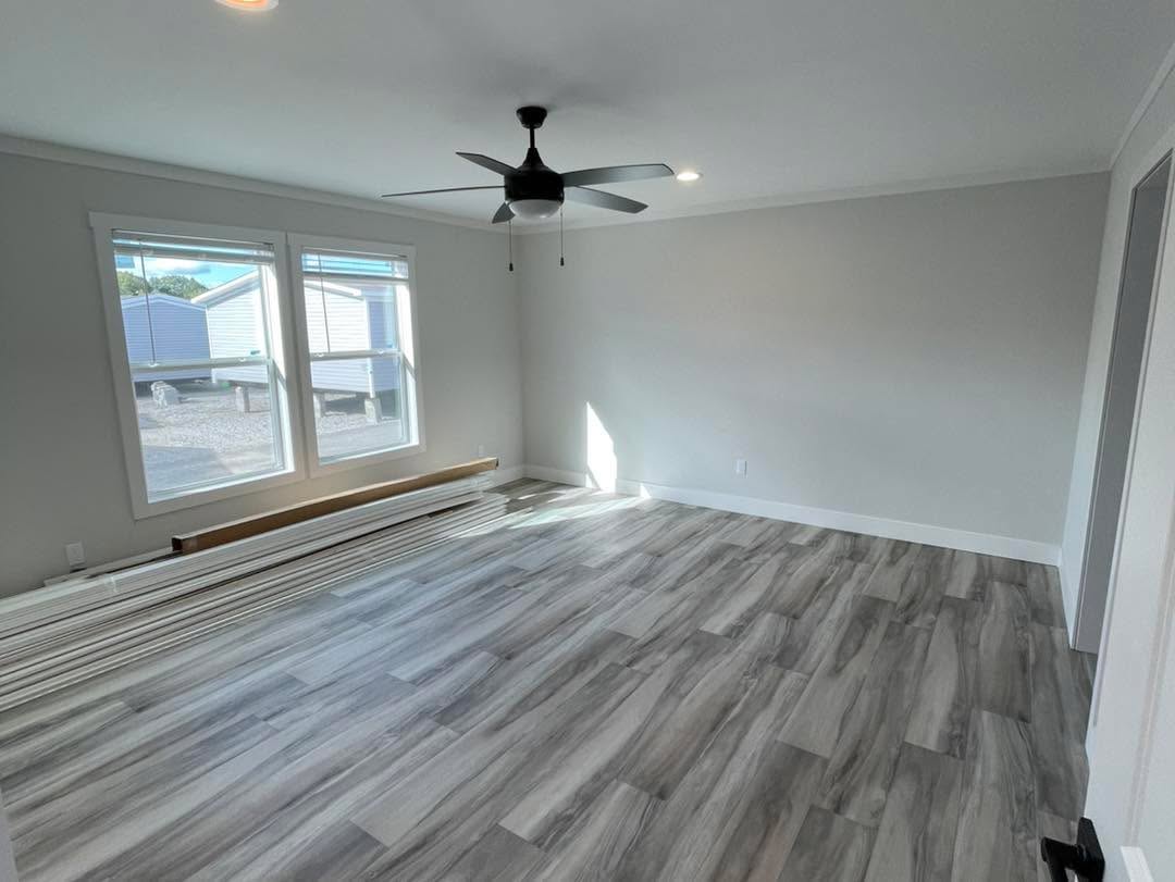 A bright, empty room with gray wood flooring, light gray walls, a ceiling fan, large windows allowing natural light, and stacked floorboards.