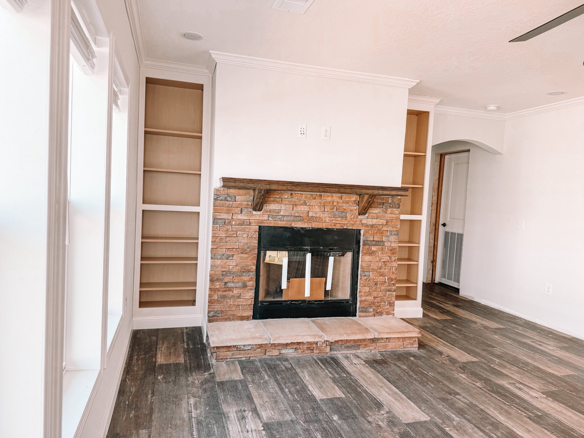 A cozy living room features a stone fireplace centered between two wooden shelves. The room has white walls and rustic wood flooring.