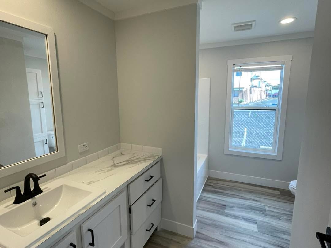 Modern bathroom with gray walls, a white marble countertop, black faucet, vanity mirror, and wood-patterned floor. Natural light through a large window.