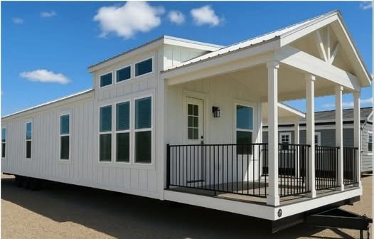 A modern, white mobile home with large windows and a small porch under a clear blue sky. The home has a gabled roof and black railings.
