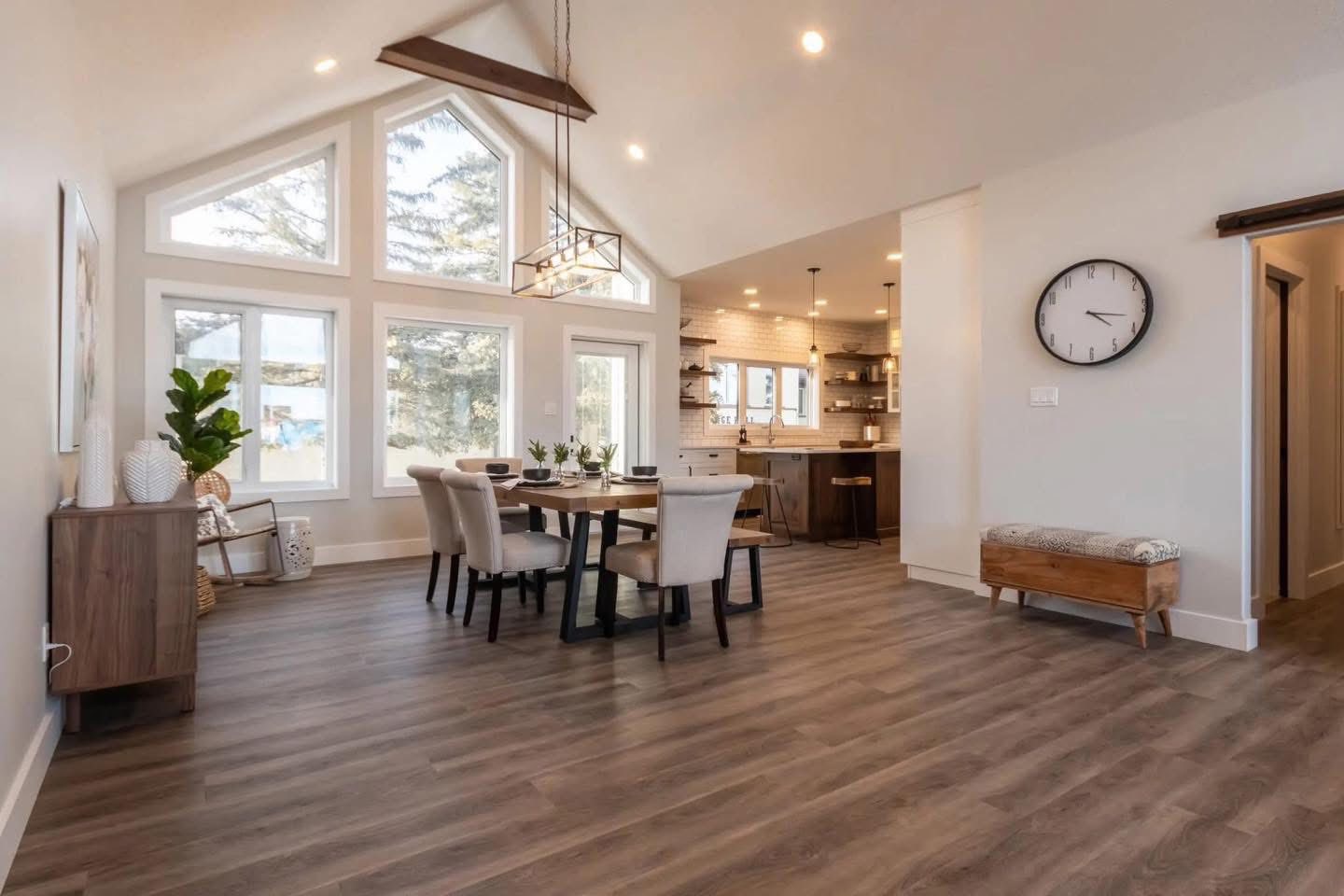 Spacious dining room with high ceilings and large windows, featuring a wooden table, beige chairs, a modern chandelier, and an open kitchen view.